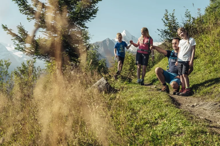 Familie beim Wandern auf einem Almweg mit Blick auf den verschneiten Berggipfel des Kitzsteinhorn im Hintergrund - Familienurlaub im Sommer in Kaprun.