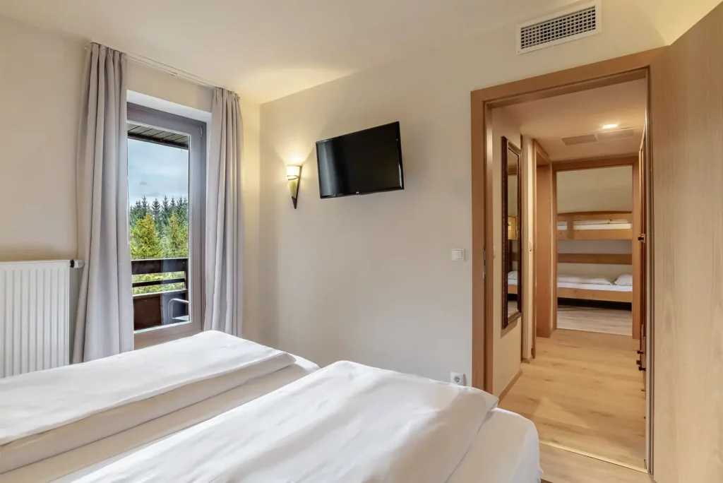 A hotel room with a double bed and a view of the balcony and the mountain scenery. A TV hangs on the wall, and through the open door you can see an adjoining room with a bunk bed.