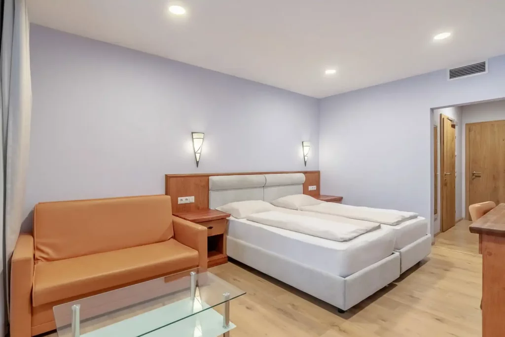 A bright hotel room with a double bed and an upholstered, light gray headboard. Next to the bed are two small wooden nightstands with wall sconces above them. On the left side of the room, an orange sofa stands in front of a small glass table. The floor is made of light wood, and the walls are painted a soft shade of lilac. In the background, an open entryway with wooden doors is visible.