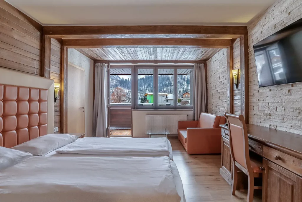 A hotel room with a double bed, wood and stone walls, and cozy lighting. To the right is a desk with a chair and a TV; to the left is a seating area. Through the large window and the balcony door, you can see a winter mountain landscape.