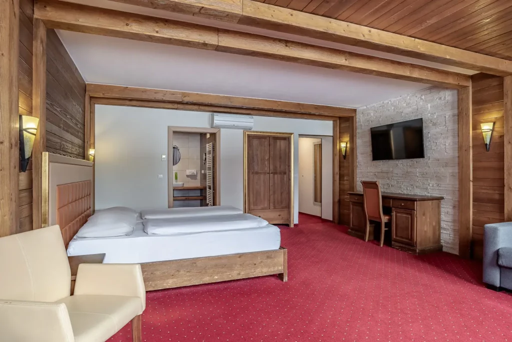 A spacious hotel room with red carpet and a double bed featuring a wooden frame and upholstered headboard, surrounded by warm wooden beams and stone wall accents. To the right are a desk with a chair and a wall-mounted TV; the bathroom and entryway are visible in the background.