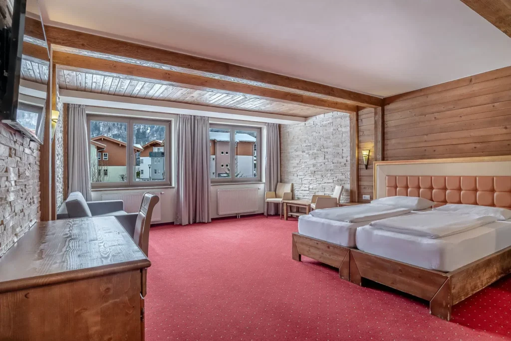 A spacious hotel room in an Alpine style, featuring plenty of wood and red carpeting. The double bed with an upholstered headboard takes center stage, complemented by a desk, seating, and light-colored stone accents on the walls. Large windows provide plenty of natural light and a view of the outdoors.
