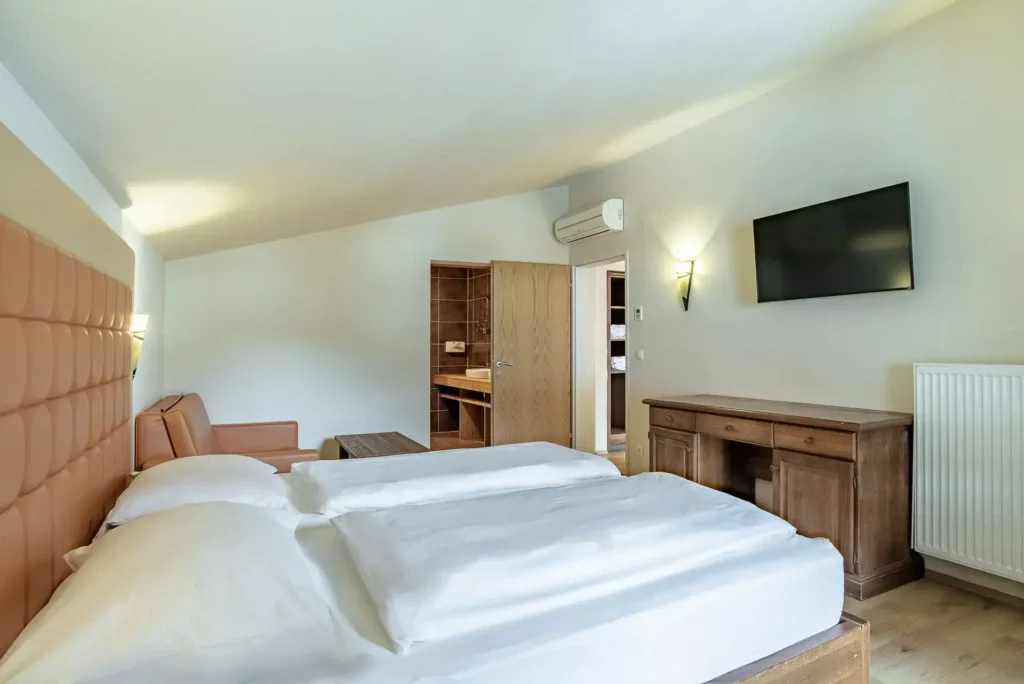 A hotel room with a double bed and an upholstered headboard. Opposite are a sideboard and a wall-mounted TV; to the left is a small seating area. The open door offers a view into the bathroom; light-colored walls and wooden accents create a warm, alpine atmosphere.