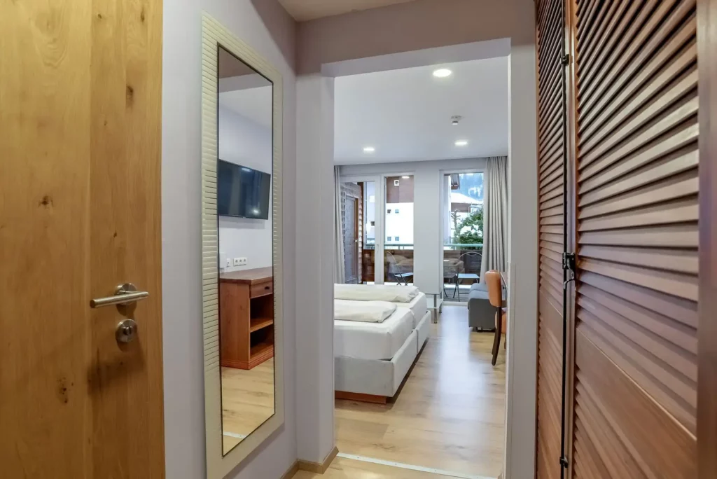 View from the entryway into a modern, bright hotel room. A large mirror and a wooden wardrobe line the hallway, which leads to the sleeping area with a double bed, a sitting area, and access to the balcony.