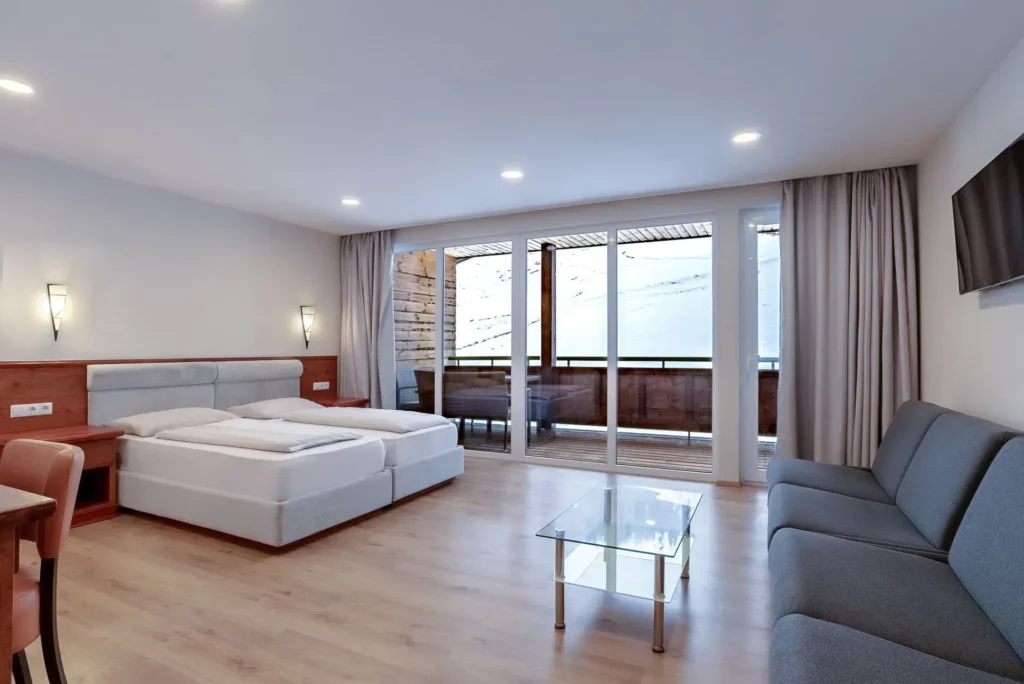 Spacious hotel room with a double bed and upholstered headboard, a comfortable sofa, and a glass coffee table. A large window with access to the balcony lets in plenty of natural light and offers a view of the snow-covered mountain landscape.