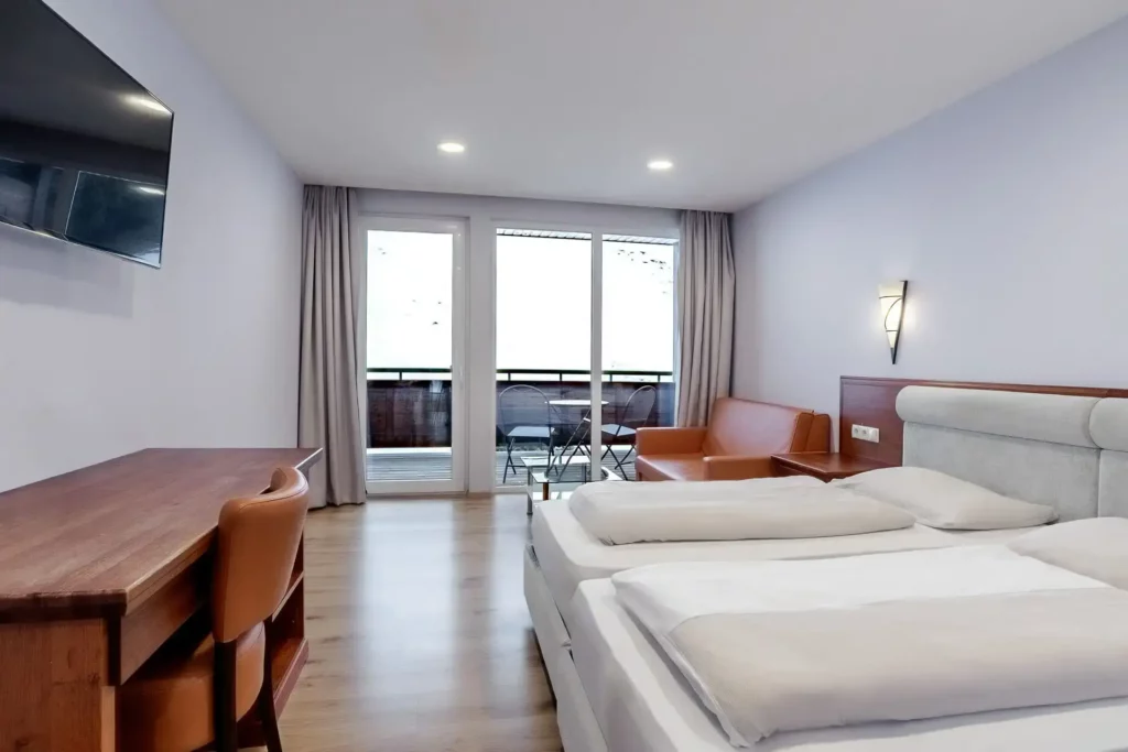 A cozy hotel room featuring a double bed, a desk, and a comfortable sitting area. Large windows leading to the balcony let in plenty of natural light and provide a space to relax while enjoying the view outside.