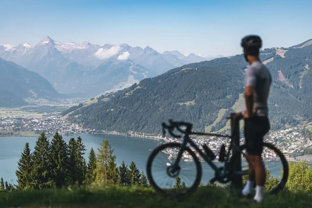 Ein Radfahrer steht mit seinem Rennrad auf einer Anhöhe und blickt über den Zeller See und die Stadt Zell am See. Im Hintergrund erheben sich die Alpen mit dem schneebedeckten Kitzsteinhorn unter blauem Himmel.